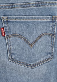 Buzunar posterior din denim albastru deschis, cu un design distinct de cusături curbate. Prezintă o etichetă roșie cu marcă pe cusătura stângă.