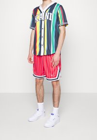 Gestreept shirt met korte mouwen in blauw, groen, geel en rood met knoopsluiting, gecombineerd met rood gestreepte shorts en witte sneakers.