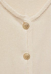 Nahaufnahme einer cremfarbenen Strickjacke mit strukturierten Kanten und zwei verzierten goldenen Knöpfen entlang der vorderen Öffnung.