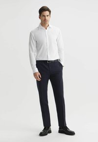 Biała, długosleeve koszula z guzikami włożona w ciemnoniebieskie, dopasowane spodnie, w połączeniu z czarnymi skórzanymi butami i czarnym paskiem.