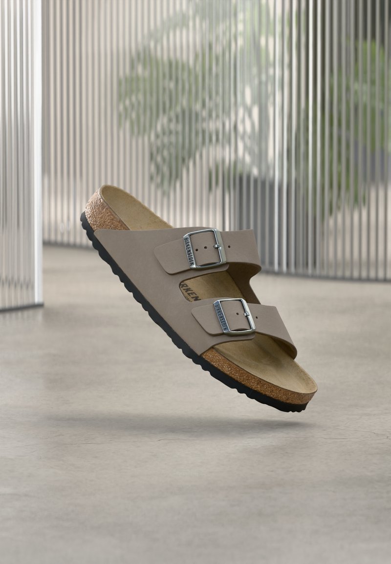 Σανδάλι Birkenstock σε χρώμα ταουπέ με δύο ρυθμιζόμενες μεταλλικές αγκράφες, ανατομική φάσα φελλού και μαύρη σόλα, αιωρούμενο πάνω από λεία επιφάνεια με θολό φόντο.