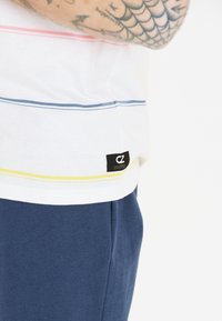 Valkoinen lyhythihainen paita, jossa on sinisiä, vaaleanpunaisia ja keltaisia vaakasuoria raitoja; musta tuotemerkki etiketti sivussa; yhdistetty tummansinisiin shortsit.