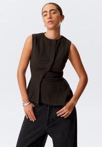 Brun senketopp med teksturert materiale, en tettsittende bodice og asymmetrisk kant, kombinert med svarte vide bukser.