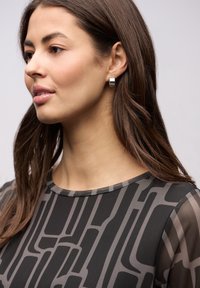 Czarny top z geometrycznymi szarymi wzorami, okrągłym dekoltem i przezroczystymi rękawami. Srebrna kolczyk w kształcie koła dodaje metalicznego akcentu. Gładka faktura.