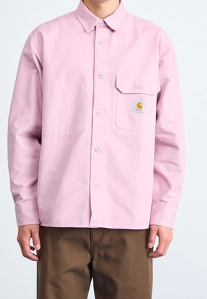 Homme portant une chemise rose clair boutonnée avec une poche avant à gauche et un pantalon marron, debout devant un fond uni.