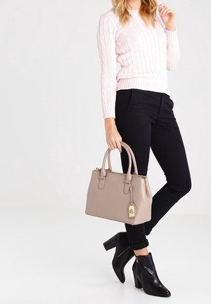 Mujer con un suéter de punto trenzado color rosa claro, pantalones negros, botines negros de tacón y sosteniendo un bolso beige con doble asa.