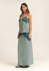 Maxikjole med figursyet overdel og udvidet nederdel i blødt grønt stof. Har en delt detalje med mørkegrønne accenter og tynde stropper.
