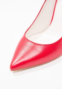 Chaussure à talon haut en cuir rouge avec un bout pointu et une texture lisse, présentant une finition brillante et un design de décolleté bas.