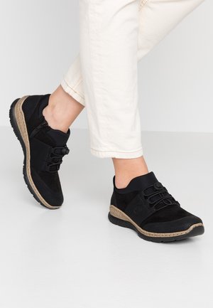 Person iført sorte sneakers med teksturerede detaljer og beige såler, kombineret med off-white bukser mod en ensfarvet baggrund.