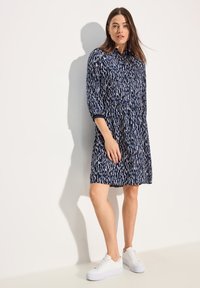 Abito camicia blu con un motivo di righe verticali bianche, chiusura con bottoni frontalmente e maniche a tre quarti, abbinato a sneakers platform bianche.