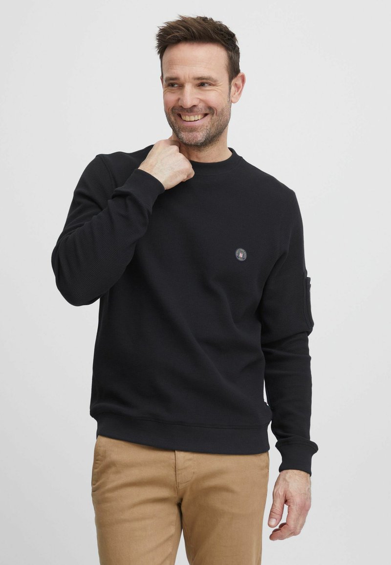 Felpa nera a collo rotondo con texture a costine, dotata di un piccolo logo sul petto e maniche lunghe. Abbinata a pantaloni color khaki.