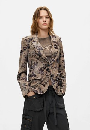 Vrouw die een blazer met bloemenpatroon draagt over een vervaagd grafisch t-shirt met zwarte cargobroek, staand met handen in de zakken tegen een witte achtergrond.