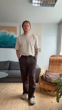 Bije kratka majica s kratkim rukavima, tamnosmeđe hlače i sive cipele na vezanje; ležeran kroj s opuštenom siluetom i minimalnim detaljima.