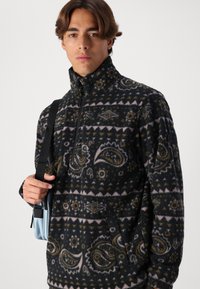 Fleece-Pullover mit Halb-Reißverschluss, schwarze Basis, Paisley- und geometrische Muster in Gelb und Pink, strukturierte Oberfläche, Fronttasche.