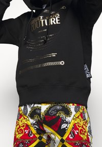 Black hoodie with gold print text and decorative accents. Paired with tapered pants featuring a colorful baroque pattern in red, black, and gold.