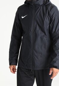 Muž v černé mikině s kapucí od Nike se zipem vpředu, dlouhými rukávy a bílým logem Nike na levé hrudi.