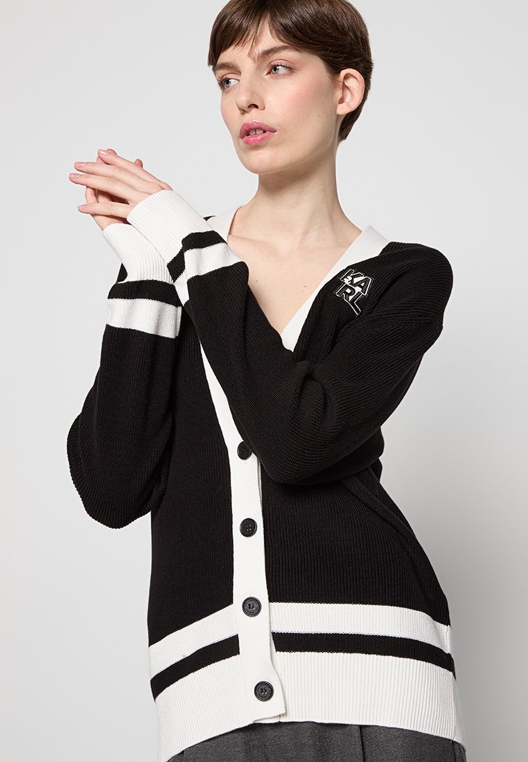 Femme aux cheveux bruns courts portant un cardigan noir et blanc à boutons, les mains jointes près du visage, regardant sur le côté devant un fond uni.