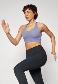 Lilla sports-bh med tynne stropper og logo i kanten, kombinert med høyt midje svarte leggings med sidelommer og jevn tekstur.