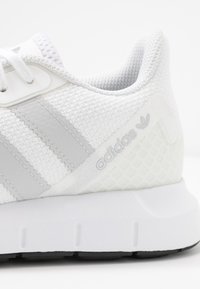 Bílá sportovní bota s texturovaným svrchním dílem, šedým designem tří pruhů a polstrovanou podrážkou. Na boku je vyražené logo.