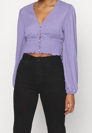 Kobieta ubrana w lawendową krótka bluzkę z długimi bufkami na rękawach, zapinaną na guziki z marszczonym pasem w talii, w parze z czarnymi spodniami z wysokim stanem.