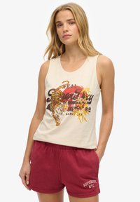 Vrouw draagt een mouwloze beige top met een kleurrijk tijger- en tekstontwerp, gecombineerd met bordeauxrode Superdry NYC short, staat met haar hand in de zak.