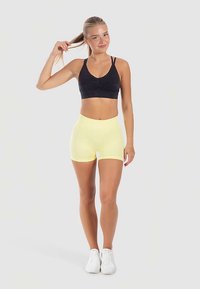 Zwarte sportbeha met dunne bandjes en plooien aan de voorkant; gecombineerd met gele sportshorts. Model draagt witte sneakers.