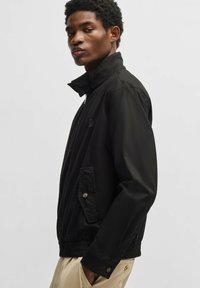 Schwarze Bomberjacke mit hohem Kragen, Reißverschluss und seitlichen Taschen. Aus glattem Stoff mit elastischen Bündchen und Saum.