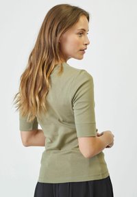 Olíva zöld, rövid ujjú póló, testhez simuló kialakítással, sima pamut anyagból, és jól látható varrással az ujjaknál és az alján.