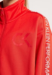 Punainen vetoketjullinen takki, joka on valmistettu teksturoidusta kankaasta. Takissa on kohokuvioitu "CK"-logo rinnassa ja valkoista tekstiä hihassa.