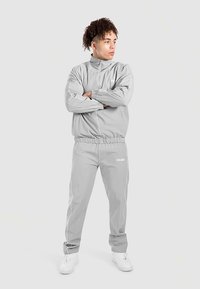 Tuta grigio chiaro in tessuto liscio, composta da giacca con zip e pantaloni con elastico in vita, rifinita con dettagli bianchi e logo.