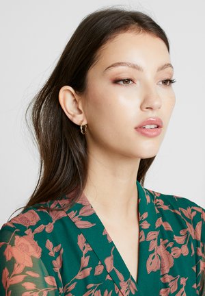 Floral print top in green with sheer sleeves, featuring a V-neckline. Gold hoop earrings accent the look, complementing the soft colors.