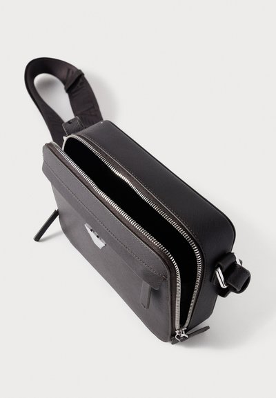 Μαύρο τσαντάκι ώμου από δέρμα με δύο θήκες που κλείνουν με φερμουάρ, υφή επιφάνειας, και ρυθμιζόμενο λουρί. Διαθέτει μπροστινή τσέπη με μεταλλικά στοιχεία.