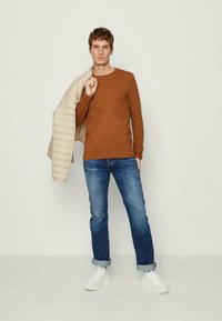 Braunes Langarmhemd, überlagert mit einer hellbeigen Steppjacke, kombiniert mit blauen Jeans und weißen Sneakern. Minimalistischer Stil.