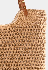 Beżowa torba na szydełku z obszernym otwartym dnem, ozdobiona plecionym wzorem oraz beżowymi skórzanymi uchwytami z mosiężnymi akcentami.