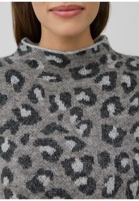 Vrouw in een fuzzy grijze trui met een zwart-witte luipaardprint en een hoge halslijn.