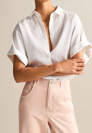 Osoba ubrana w białą, krótkorękawową koszulę z podwiniętymi rękawami i jasnoróżowe, wysokowaistowe spodnie, z założonymi skrzyżowanymi rękami.