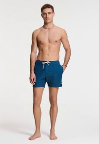 Sinised ujumispüksid tekstuurse mustriga, millel on nöörvöö ja küljele kantud logo. Modell seisab paljajalu, paljastatud ülakehaga.