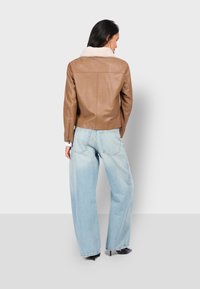 Vrouw in een tan leren jas met een witte schapenvacht kraag, lichtblauwe wijde jeans en zwarte puntige hakken, staand met de rug naar ons toe.