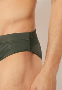 Dunkelgrüne Schwimmshorts aus glattem Stoff, mit niedrigem Schnitt und dezenter Logodetail auf der Seite. Strukturiere Oberfläche ohne Muster.