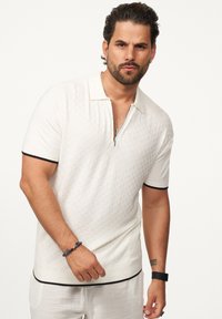 Weißes, strukturiertes Poloshirt mit einem Halbreißverschluss-Kragen, schwarzen Akzenten an Ärmeln und Saum. Kombiniert mit hellen Hosen und einem Perlenarmband.