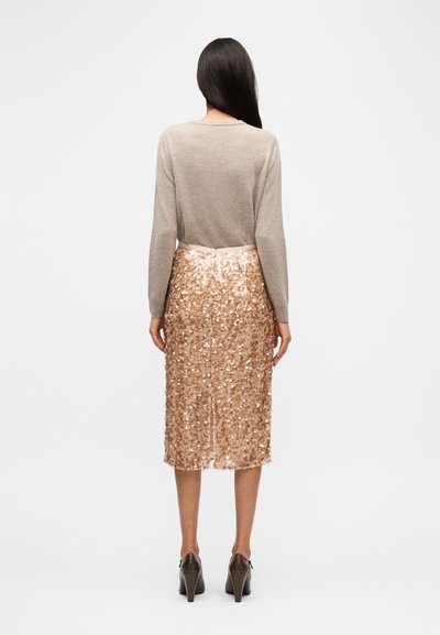 Ανοιχτό καφέ πουλόβερ με μακριά μανίκια, συνδυασμένο με φούστα μήκους μέχρι το γόνατο, με γυαλιστερές παγιέτες σε ροζ χρυσό χρώμα και σκούρα ψηλοτάκουνα παπούτσια. Ολική λεία υφή.