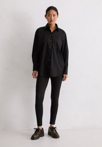 Jonge vrouw die tegen een witte muur staat, gekleed in een oversized zwart overhemd met knopen, strakke zwarte broek en donkerbruine veterschoenen.