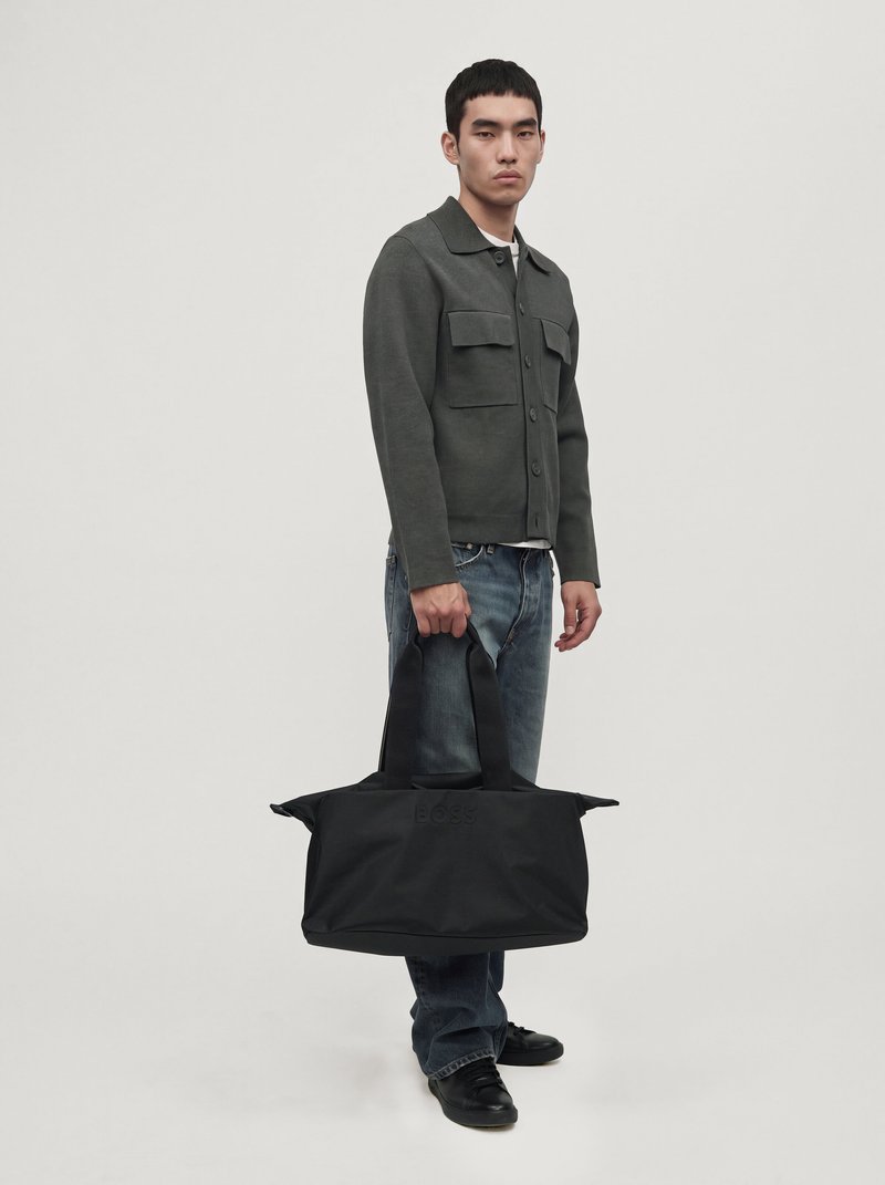 Un homme porte une veste gris foncé et un jean, et tient un grand sac de voyage noir sur un fond clair uni.