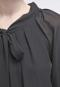 Svart genomskinlig blus med knutband vid halsen, samlad tyg vid axlarna och en genomskinlig ärm på höger sida.