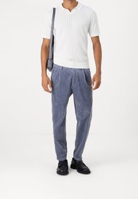 Bílá žebrovaná polokošile s krátkým rukávem a výstřihem do V, v kombinaci s modrými manšestrovými kalhotami s plisováním a černými botami, doplněná kabelkou přes rameno.