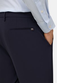 Granatowe spodnie o gładkiej fakturze, z zamkiem błyskawicznym w tylnej kieszeni i małym akcentem w postaci logo. W komplecie z jasnoniebieską koszulą w paski.