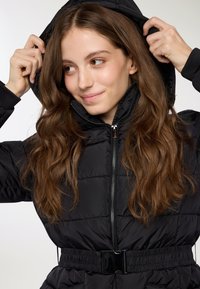 Czarna puchowa kurtka z wysokim kołnierzem i kapturem. Posiada zamek błyskawiczny z przodu, elastyczne mankiety oraz ściągnięty pas z paskiem. Teksturowany materiał.