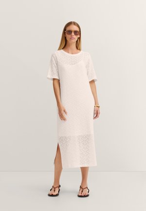 Frau trägt ein weiß gemustertes Midikleid mit kurzen Ärmeln, braune Sonnenbrille, ein goldenes Armband und dunkle geflochtene Sandalen, steht vor einem einfarbigen Hintergrund.