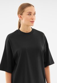 Tricou negru supradimensionat cu mâneci scurte, căzute. Confecționat dintr-un material moale din bumbac și are un guler rotund.