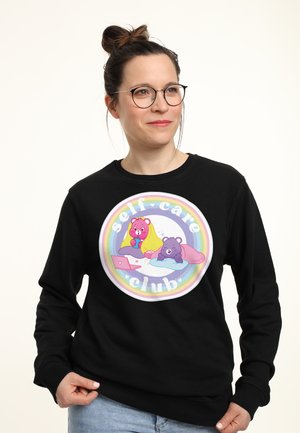 Žena s brýlemi nosící černou mikinu s obrázkem dvou kreslených medvědů, notebookem a textem „self care club“ uvnitř duhového kruhu.